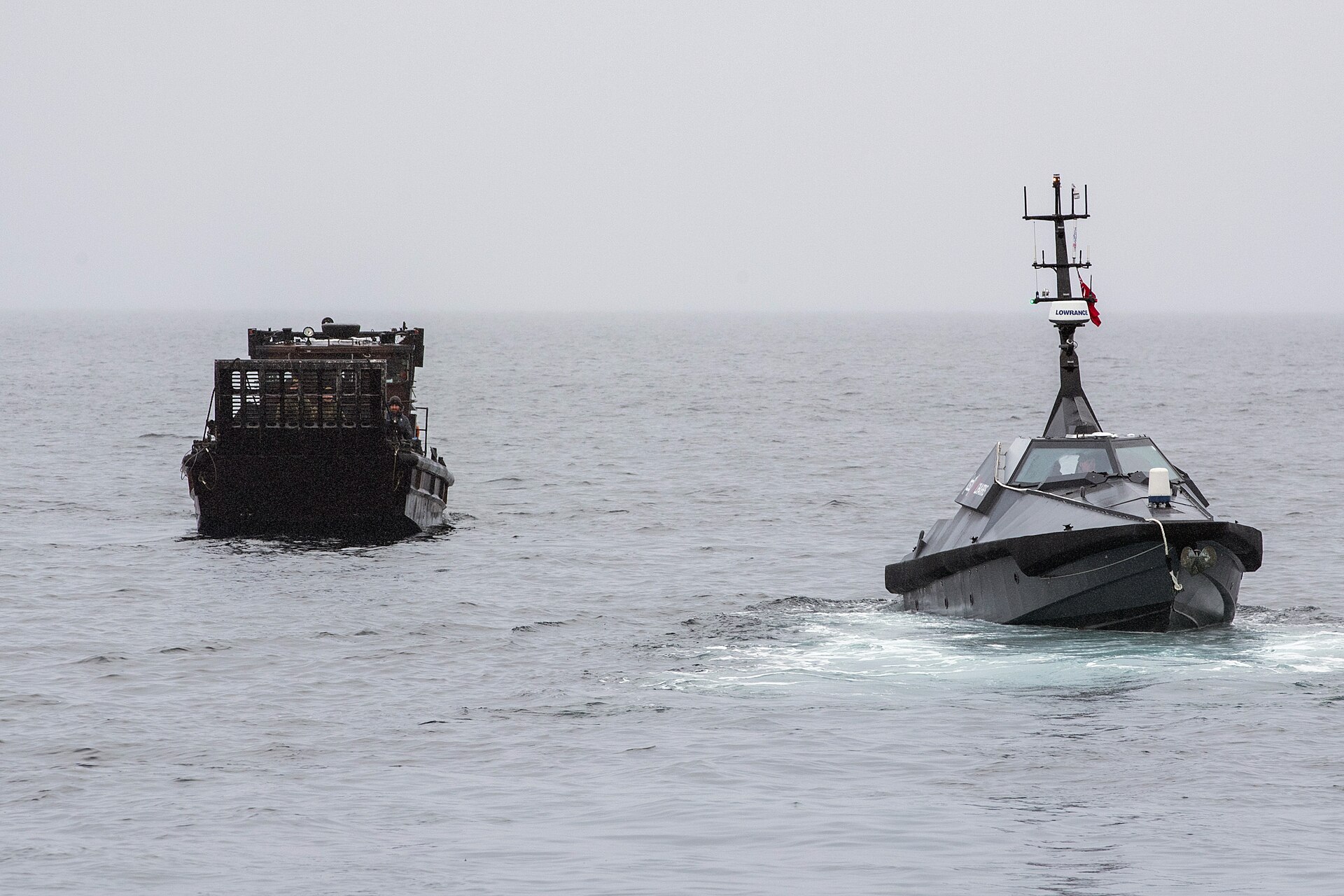 An uncrewed surface vessel with a mast and radar approaching at sea, with a Royal Marines landing craft behind it